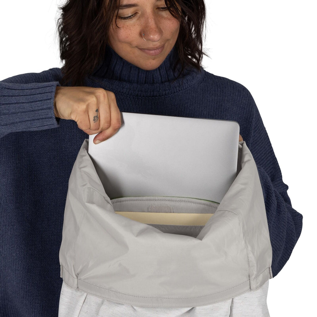 The image shows a person wearing a dark blue, ribbed long-sleeve sweater, holding a gray, flap-top backpack. They are in the process of placing or removing a silver laptop from the main compartment of the backpack, which appears to have a light-colored in