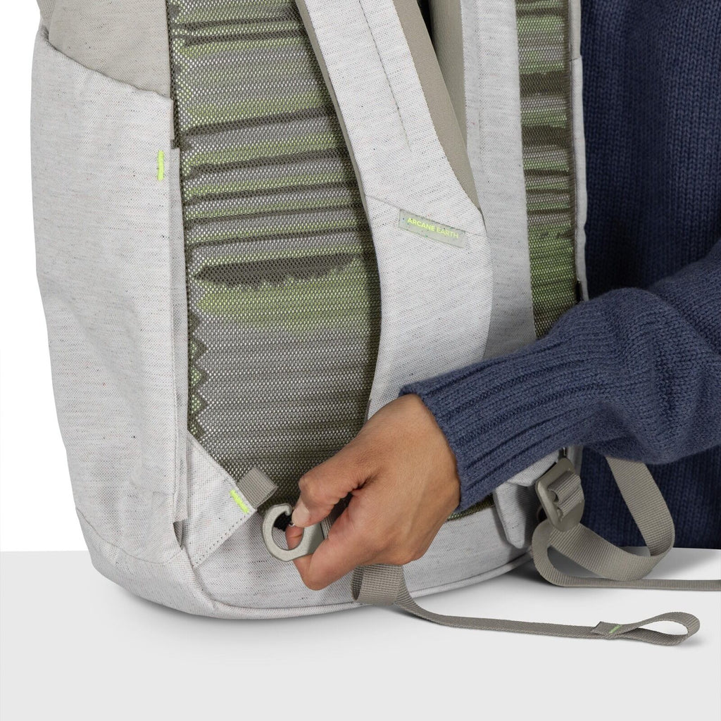 The image shows a close-up of a person adjusting or pulling a strap on a light gray backpack. The backpack features green patterned fabric visible between the padded sections of the back panel. The person is wearing a dark blue knitted sweater, and their