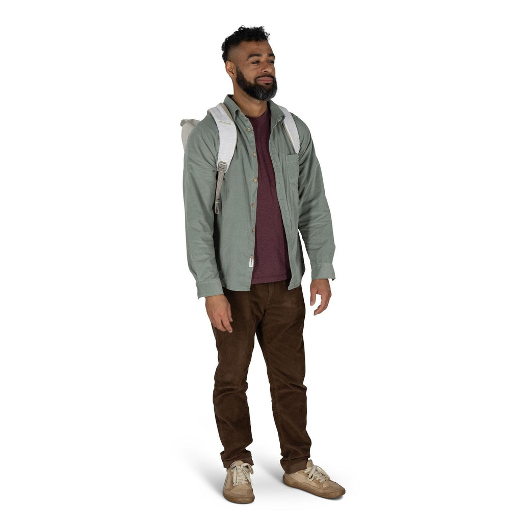 The image shows a person standing, dressed in casual attire. They are wearing a gray backpack on their shoulders. The individual has on a green button-up shirt layered over a burgundy shirt, paired with brown pants. Completing the outfit are beige sneaker