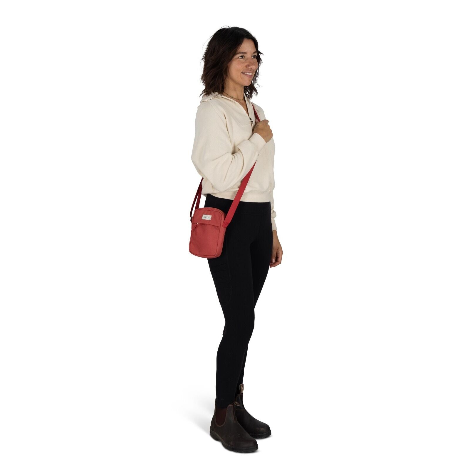 The image features a woman standing against a plain white background. She is wearing a light-colored sweatshirt and black leggings, paired with dark ankle boots. Draped across her body is a small red crossbody bag, which she holds with one hand. Her hair