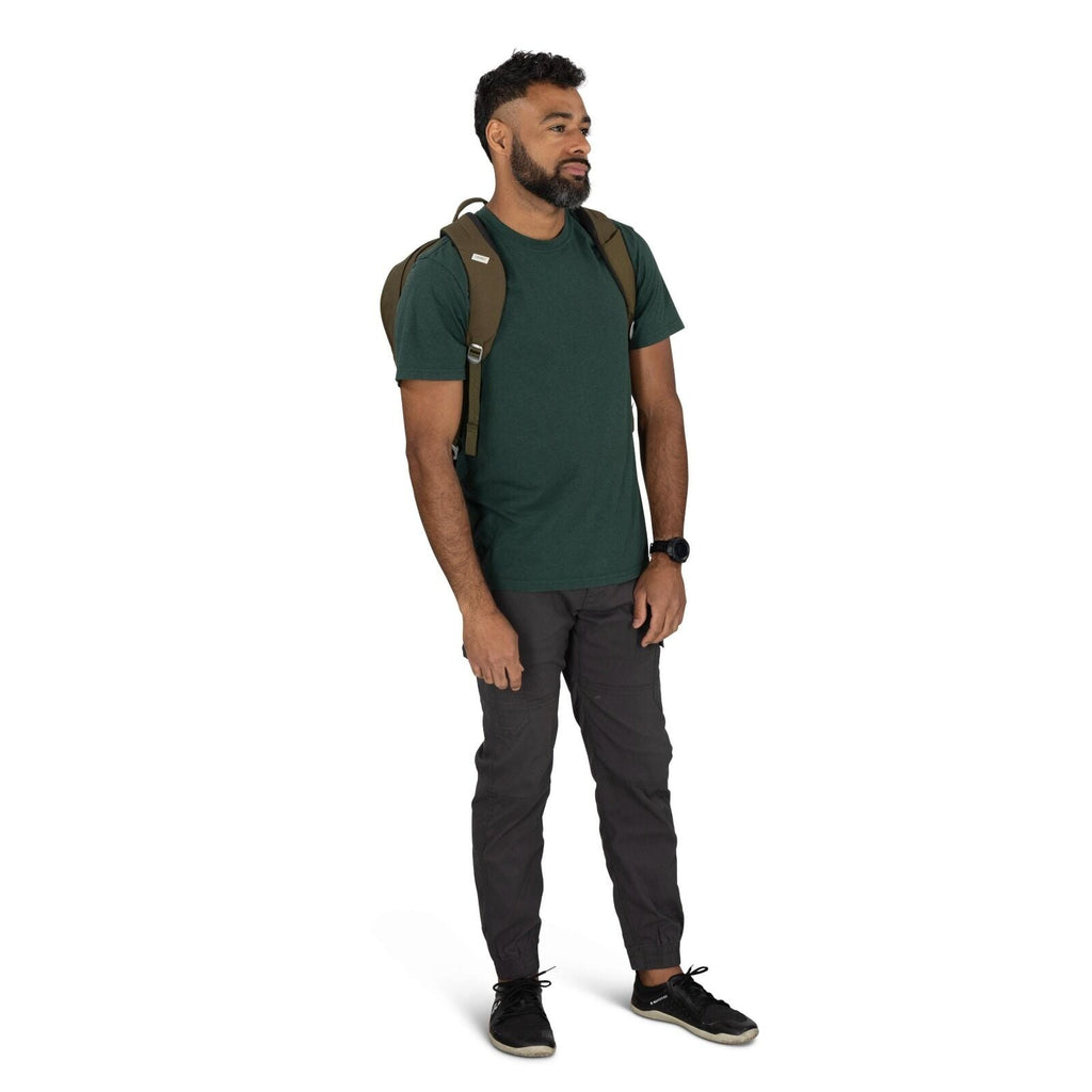 The image shows a man standing in a casual pose, wearing a dark green T-shirt and dark gray cargo pants. He has a small brown backpack on his shoulders, suggesting he might be prepared for a casual outing or a day hike. On his wrist, he is wearing a black