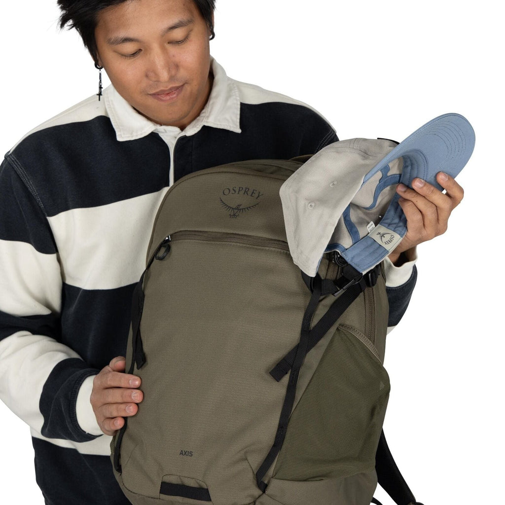 The image shows a person wearing a black and white striped long-sleeve shirt. They are holding an Osprey backpack, which is olive green with black straps, and has the "OSPREY" logo with an axis design. The person is also holding a light gray and blue base