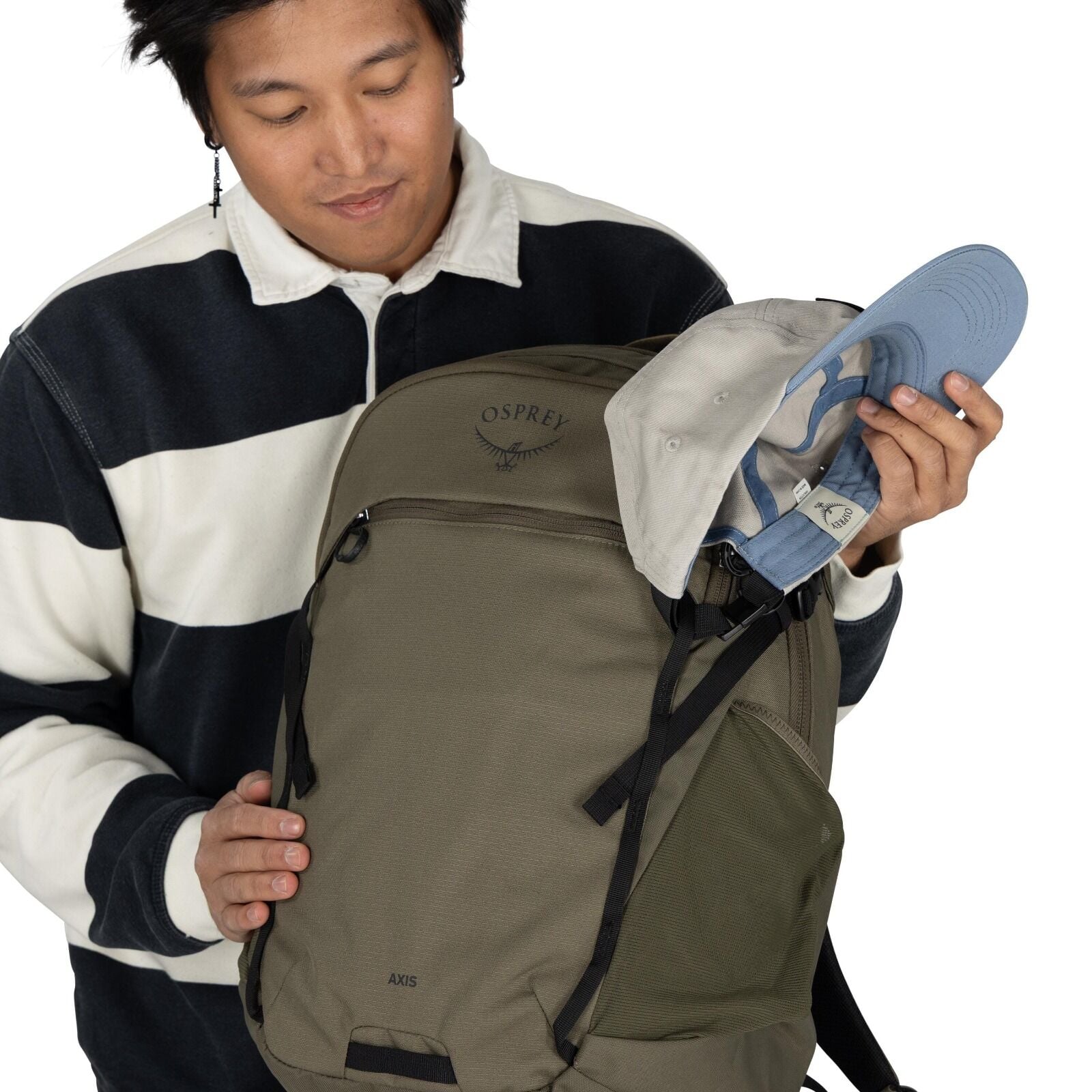 The image shows a person wearing a black and white striped long-sleeve shirt. They are holding an Osprey backpack, which is olive green with black straps, and has the "OSPREY" logo with an axis design. The person is also holding a light gray and blue base