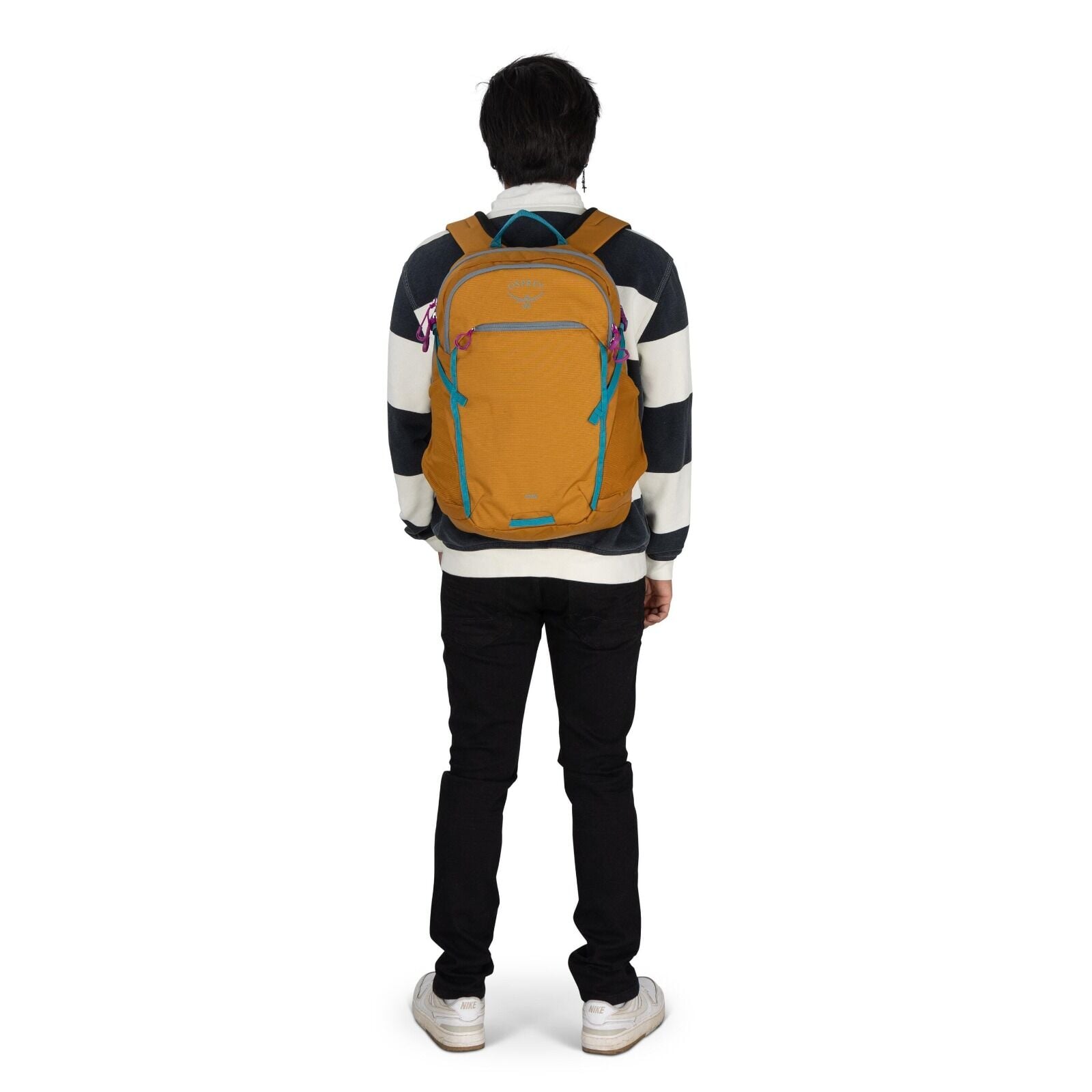 The image shows a person standing with their back to the camera, wearing a striped sweater and black pants. The individual has on a mustard-colored Osprey backpack, which features turquoise zippers and accents, along with a distinctive logo visible at the
