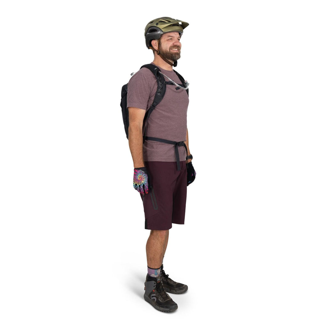 The image depicts a man dressed in outdoor gear, likely for biking or hiking activities. He is wearing a green safety helmet, a maroon short-sleeved shirt, and burgundy shorts. On his hands are colorful gloves featuring a tie-dye pattern. His feet are ado
