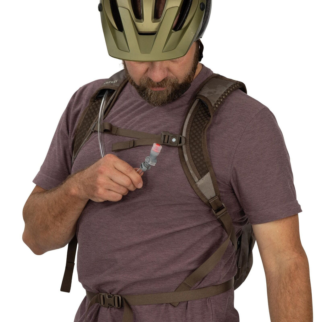 The image shows an individual wearing a light purple short-sleeved shirt and a backpack with shoulder straps and a chest buckle. The backpack is equipped with a hydration system, indicated by the clear tube that leads to a bite valve, which the person is