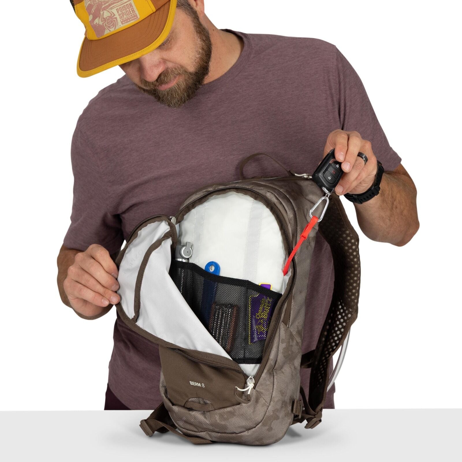 The image shows a person holding open a brown backpack, revealing its contents. The person is wearing a maroon t-shirt and a brown and yellow cap. The inside of the backpack is neatly organized with various items visible in mesh pockets, including a blue