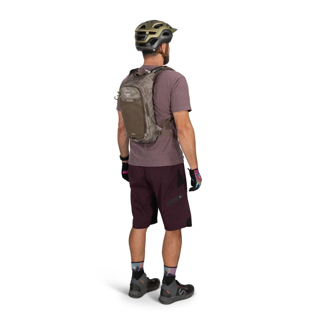The image features a man standing with his back turned, wearing a biking outfit. He is dressed in a brown cycling helmet and a purple short-sleeve shirt, accompanied by dark purple shorts with black patches. On his back, he carries an Osprey backpack, and