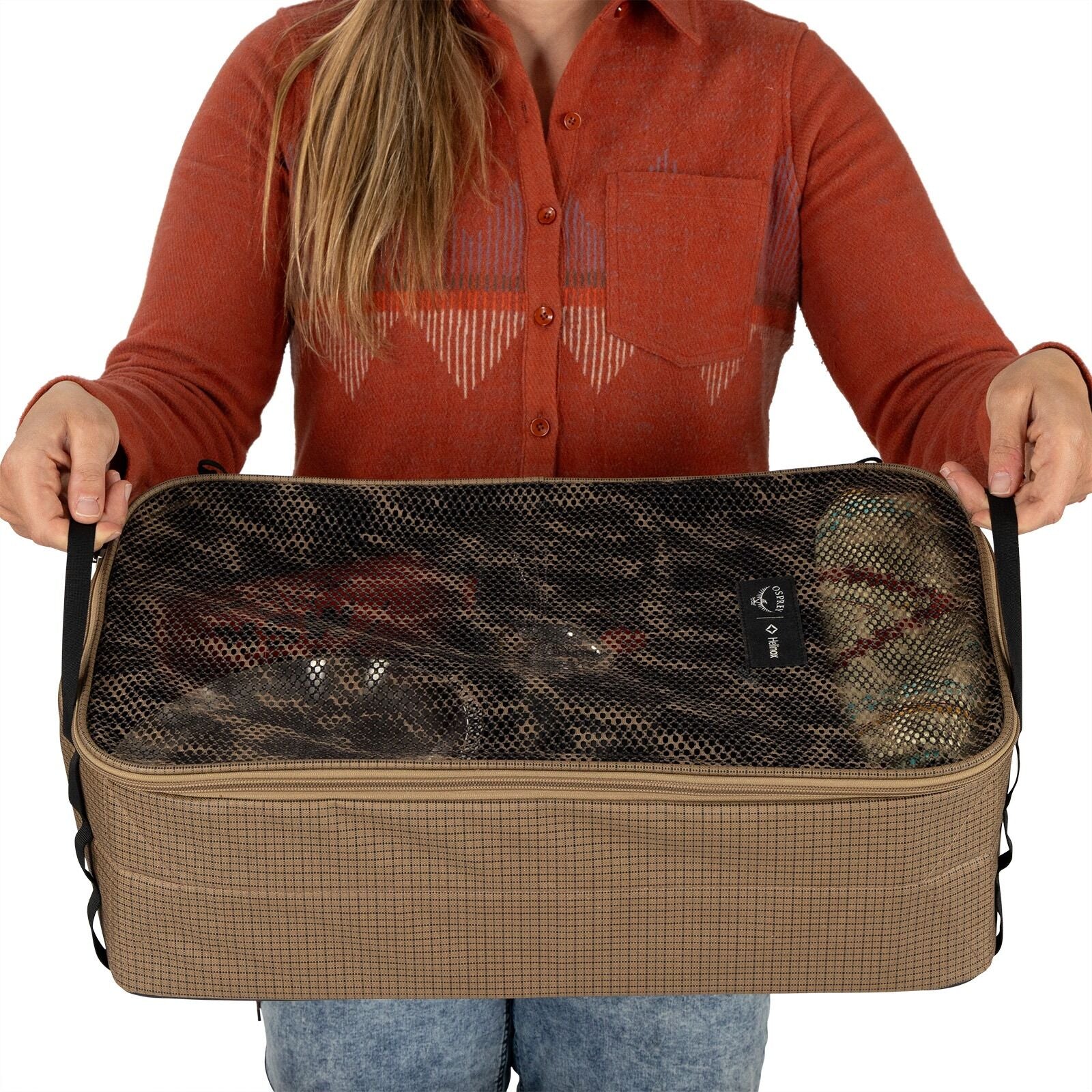 A person in an orange shirt holds a beige rectangular storage bag with a black mesh top, revealing packed items inside. The tone is practical.