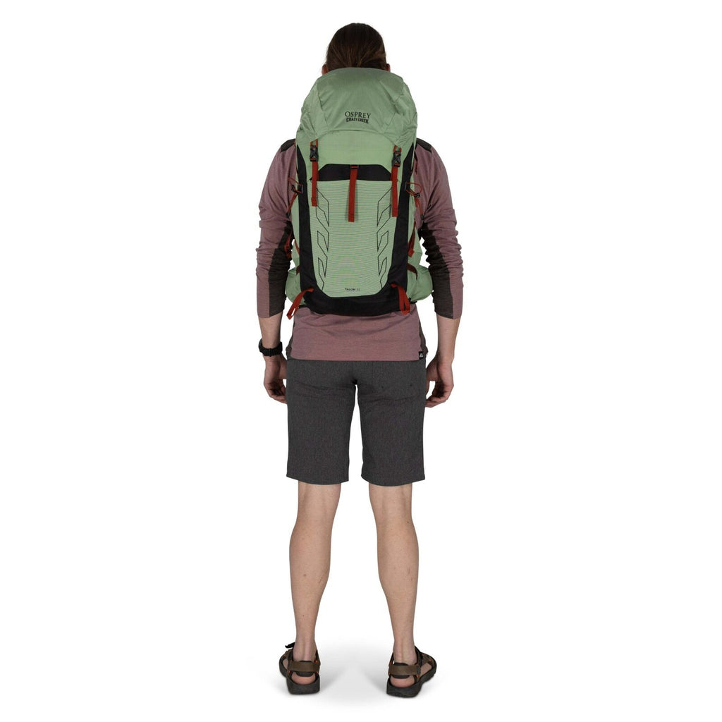 A person stands facing away, wearing a light green Osprey backpack and grey shorts, ready for an outdoor adventure.