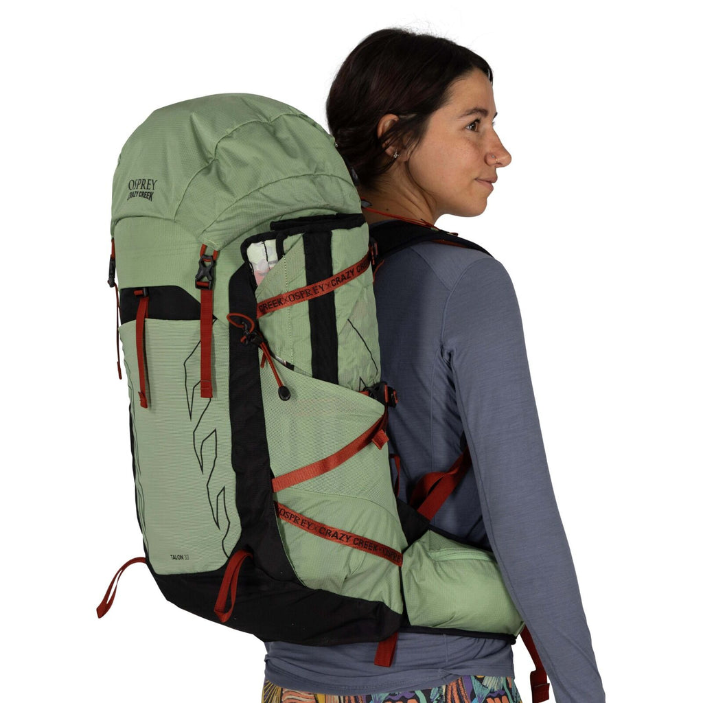A person wears a large, green and black Osprey / crazy Creek Talon 33 backpack, designed for outdoor adventures, featuring multiple pockets and straps.