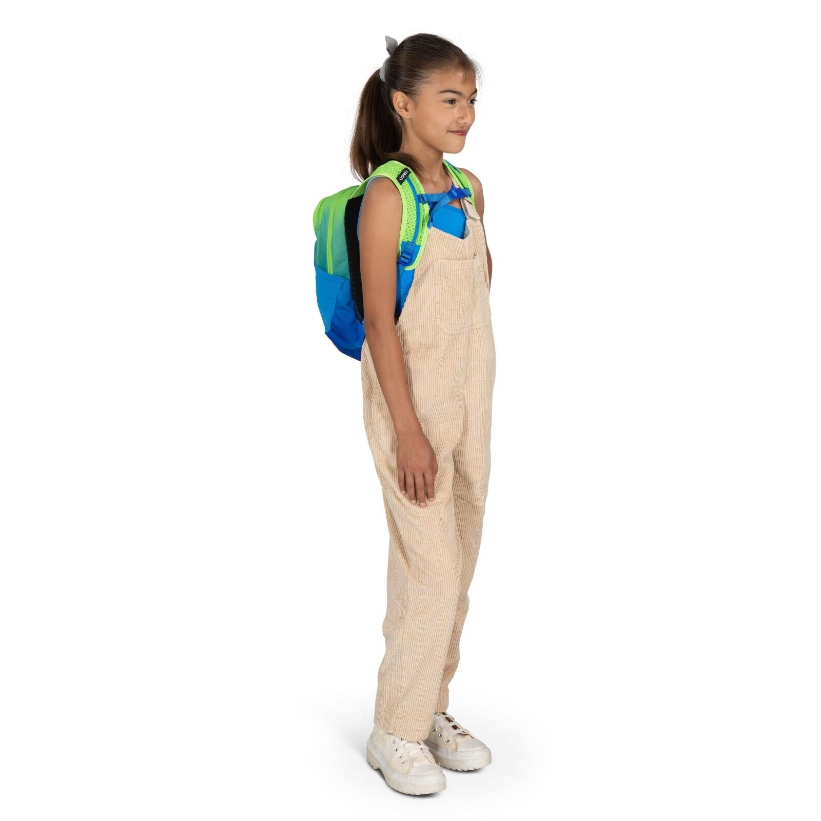 The image shows a young person standing sideways, wearing beige overalls and a sleeveless black top. They are carrying a colorful backpack, predominantly blue with green straps. The person is also wearing white sneakers. The overall look suggests casual a