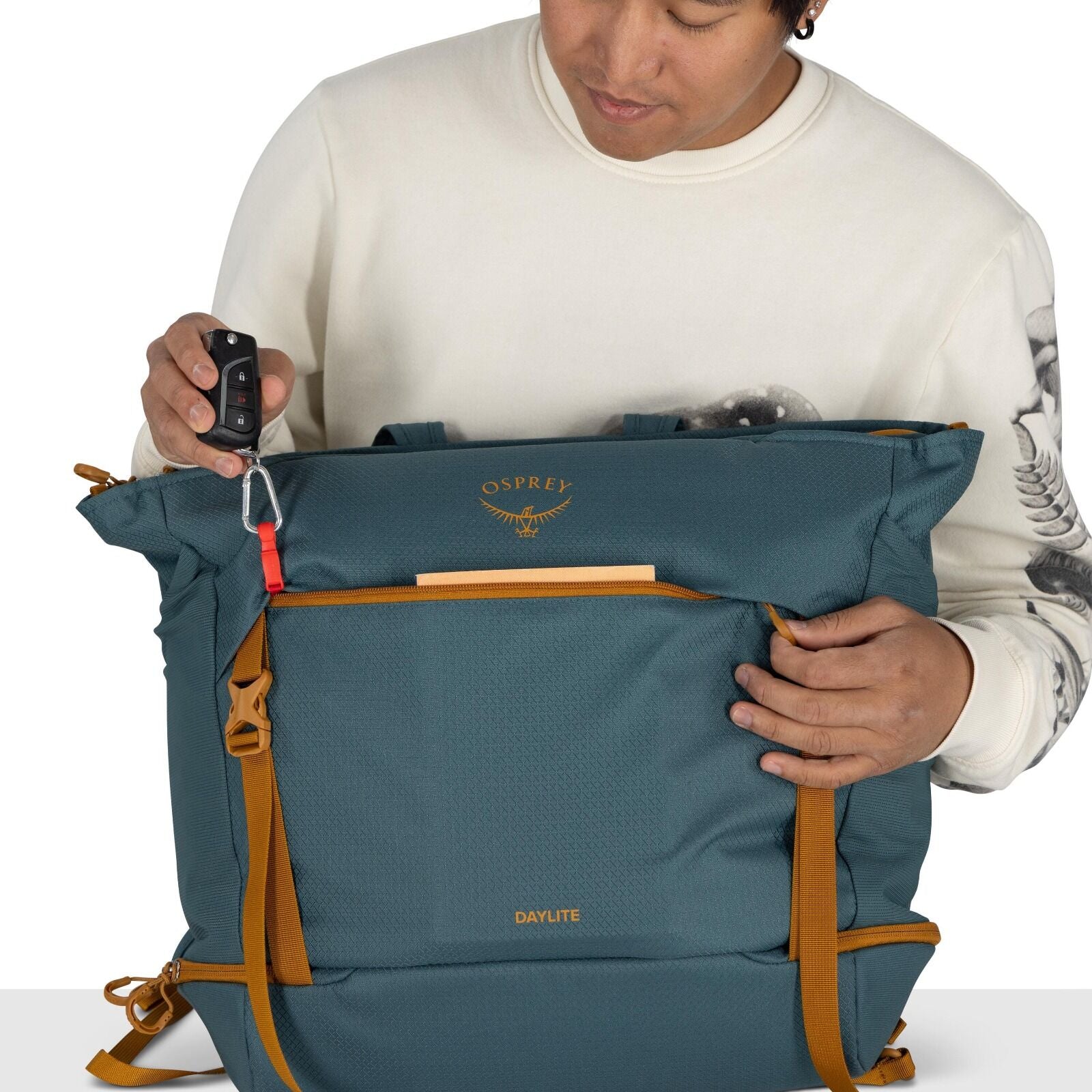 The image depicts a person wearing a white sweater with dark artistic designs on the sleeves, holding a teal-colored Osprey backpack. The backpack has distinct tan accents, including straps and zippers. The person is using a carabiner attached to the bag'