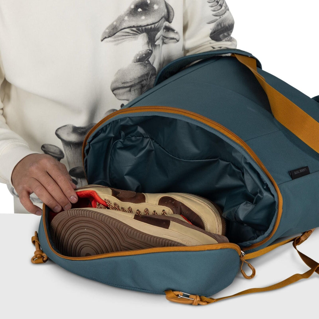 The image shows a close-up view of a person packing a pair of beige and brown sneakers into a duffel bag. The duffel bag is a deep teal color with brown straps and zippers, and has a separate shoe compartment being used. The person is wearing a cream-colo