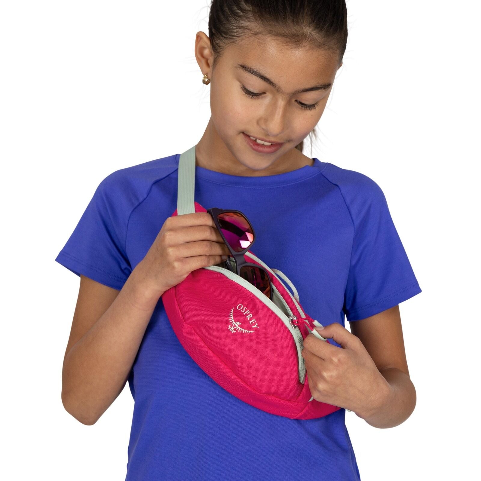 The image features a person wearing a blue short-sleeved shirt and holding a pink Osprey waist bag or fanny pack. The bag is slung across their body, with the strap going over the right shoulder. The person is in the process of placing a pair of sunglasse