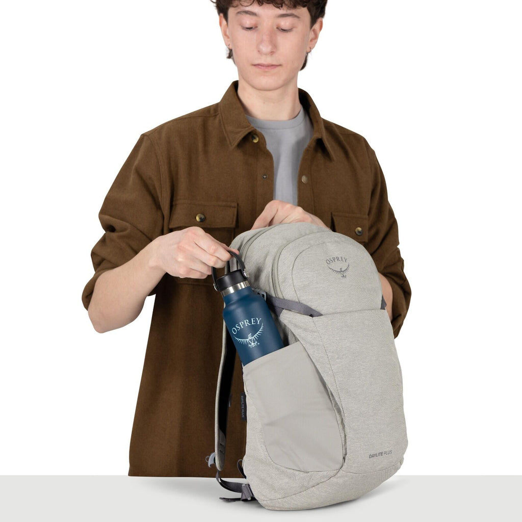 A person is placing a blue Osprey water bottle into the side pocket of a light gray Osprey backpack. The backpack has a clean, minimalist look with a front shove-it pocket and side stretch pocket. The person is wearing a brown overshirt over a gray tee. T