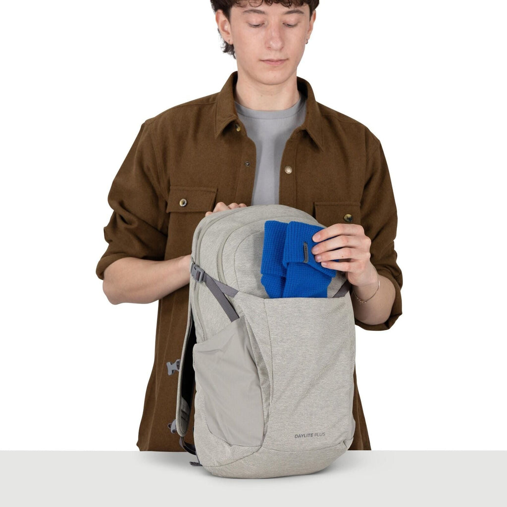 A person is holding a light gray Osprey Daylite Plus backpack and placing a folded bright blue garment into its front shove-it pocket. The backpack has a stretchy side pocket, gray compression straps, and a soft heathered fabric. The person is wearing a b
