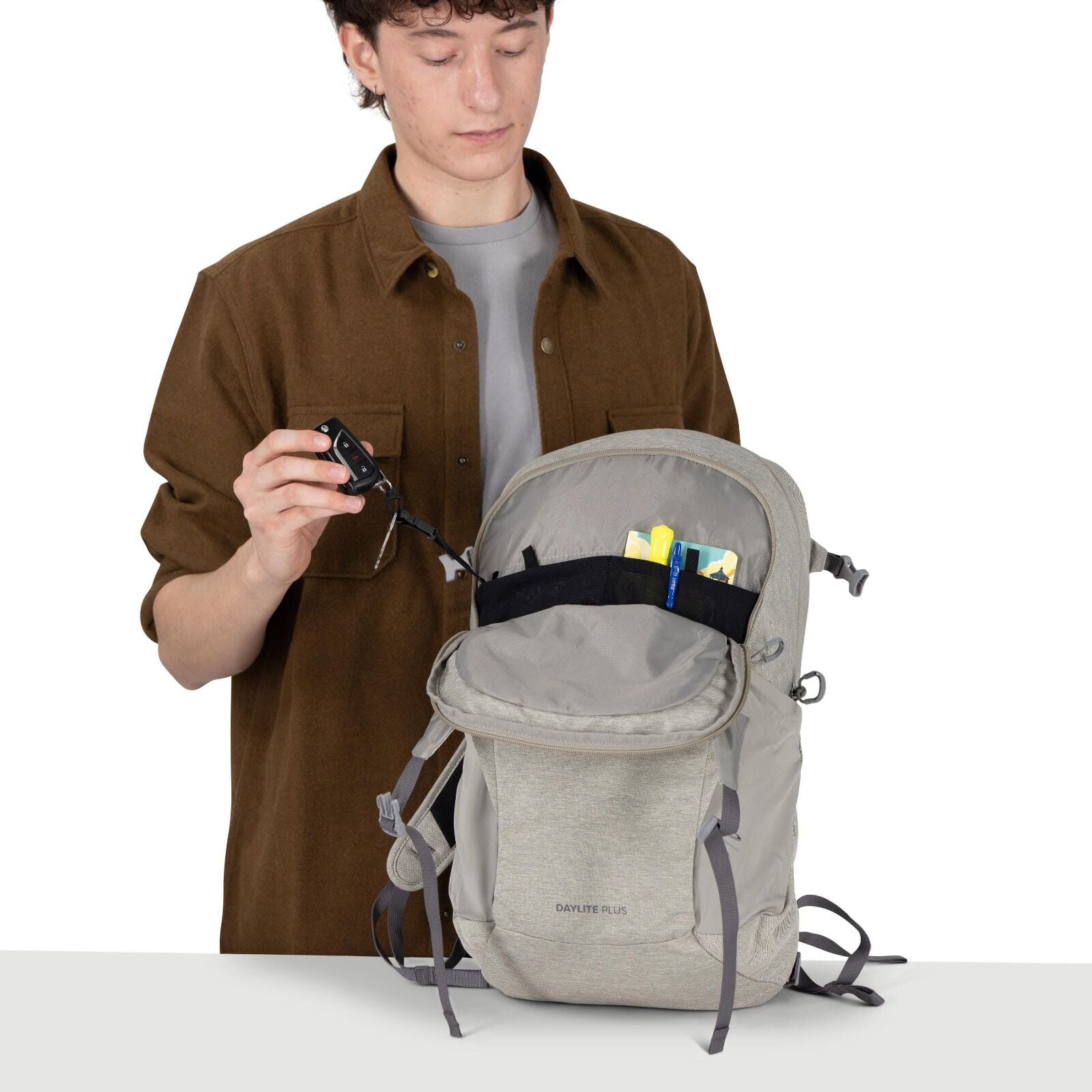 A person wearing a brown overshirt and gray T-shirt is demonstrating a light gray backpack. The bag is open, showing an organized interior with a black mesh pocket holding small items like a pen and cards. The person is pulling out a key attached to an in