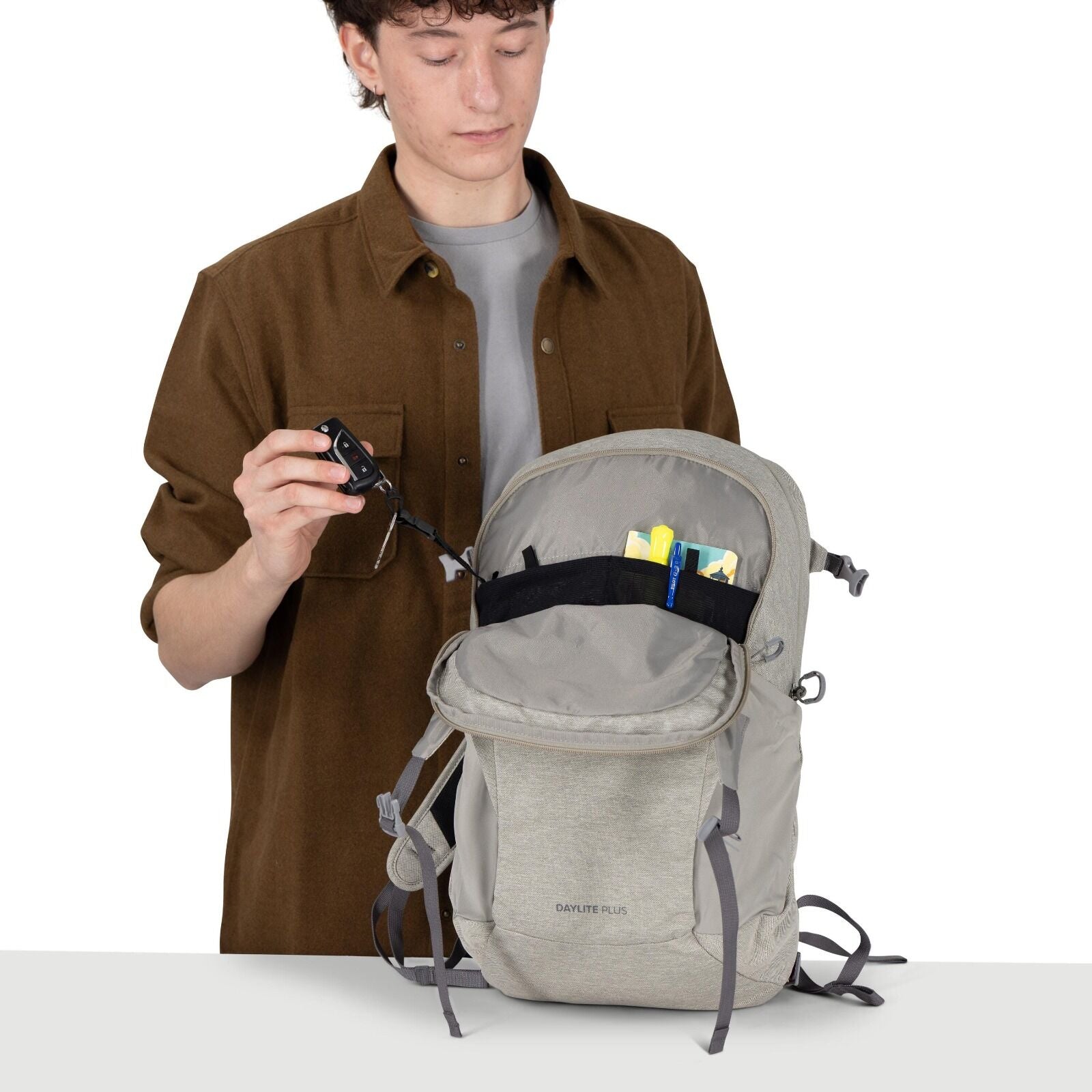 A person wearing a brown overshirt and gray T-shirt is demonstrating a light gray backpack. The bag is open, showing an organized interior with a black mesh pocket holding small items like a pen and cards. The person is pulling out a key attached to an in