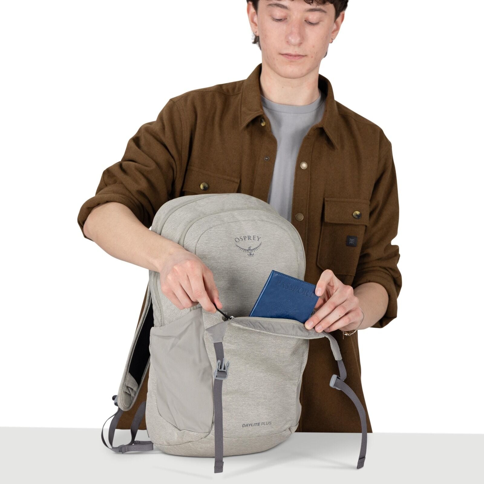 A person wearing a brown overshirt and gray tee is holding a light gray Osprey backpack. They are opening the front zip pocket and placing a blue passport inside. The backpack has the Osprey logo on the upper panel and "Daylite Plus" text near the bottom.