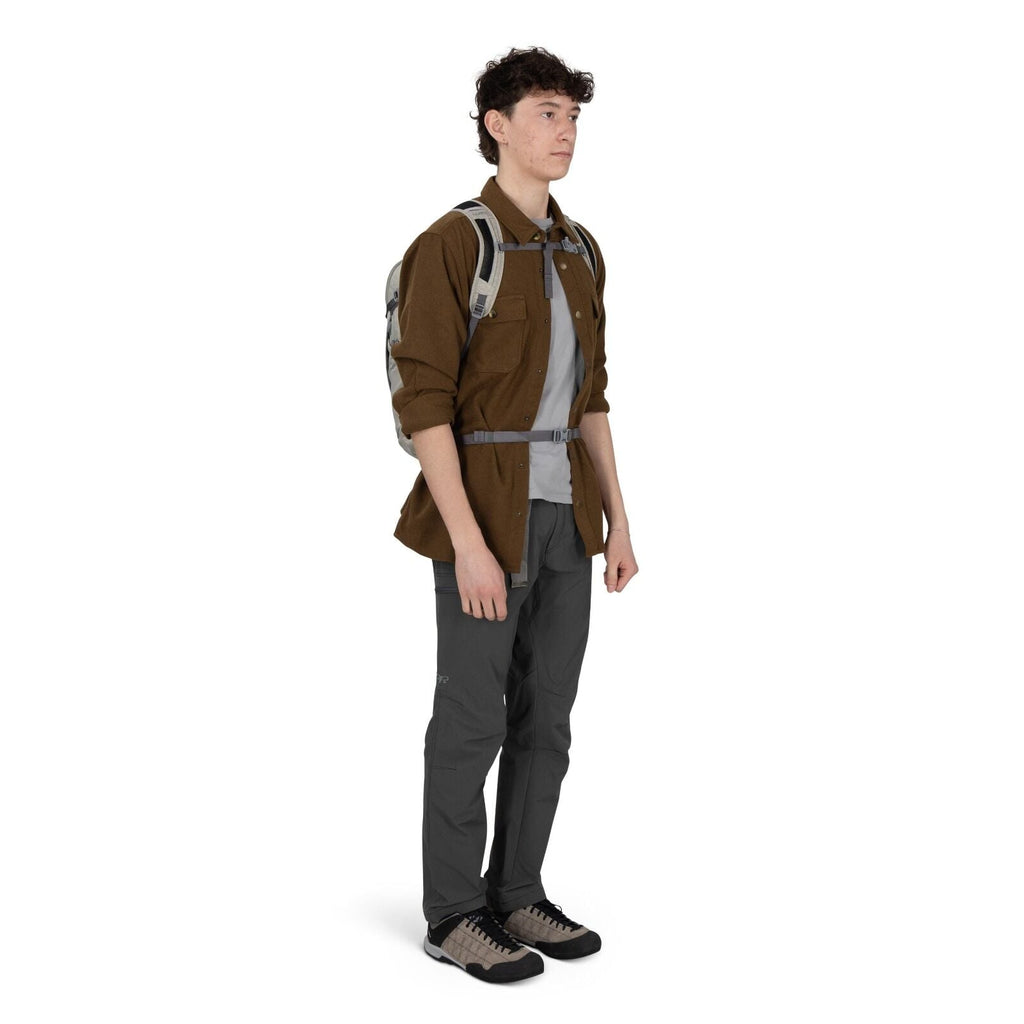 A person stands in a studio setting wearing outdoor clothing and a small backpack. They have on a brown button-up overshirt over a light gray T-shirt, charcoal-gray hiking pants, and beige-and-black trail shoes. The backpack straps are clipped across the