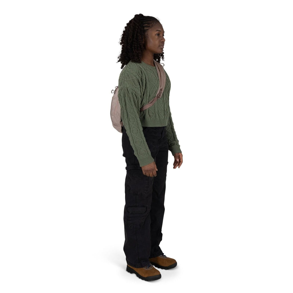 A person stands in a three-quarter view wearing a green cable-knit cropped sweater and loose black cargo pants. They have a small beige sling backpack across their chest. Brown hiking-style shoes with black soles complete the outfit. The background is pla