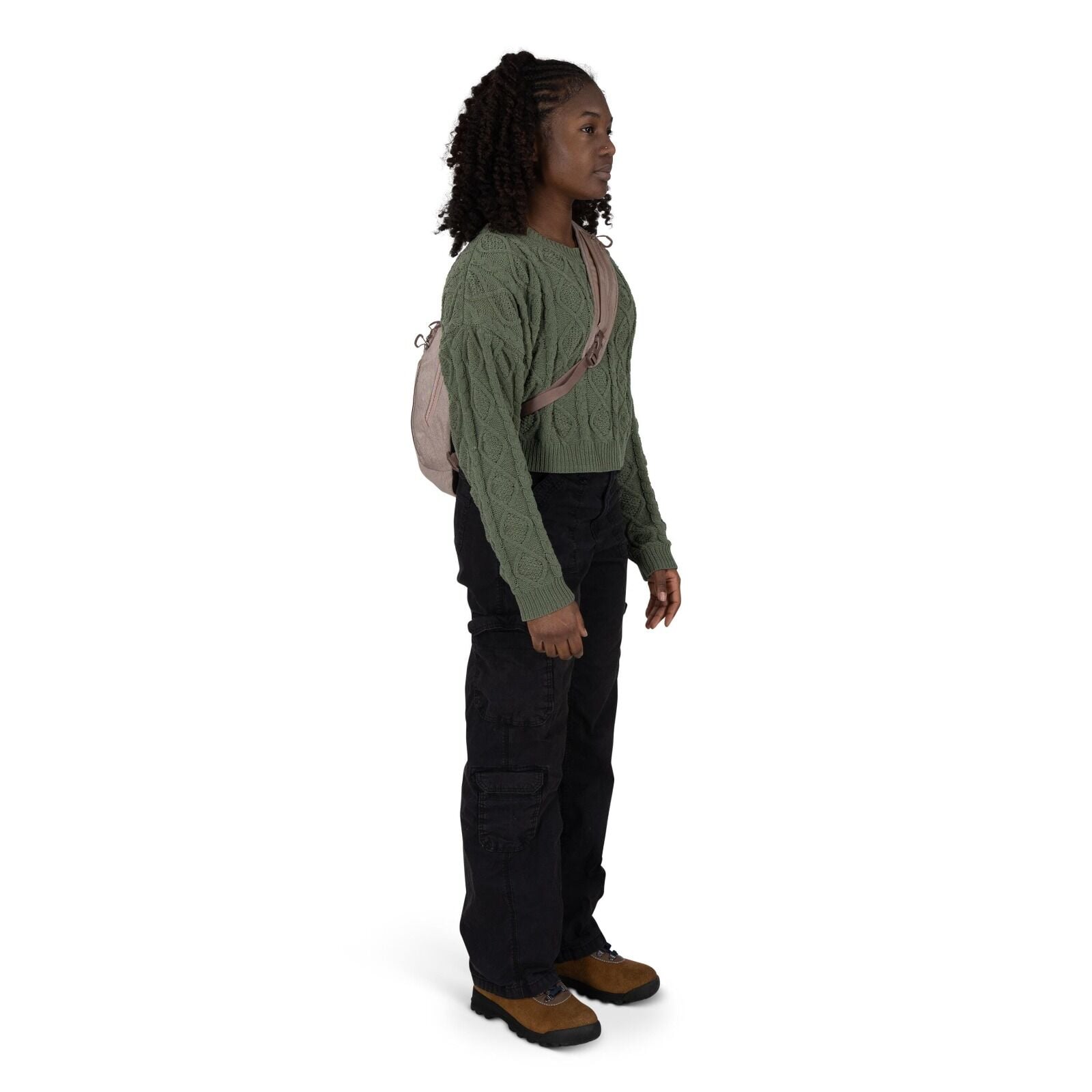 A person stands in a three-quarter view wearing a green cable-knit cropped sweater and loose black cargo pants. They have a small beige sling backpack across their chest. Brown hiking-style shoes with black soles complete the outfit. The background is pla