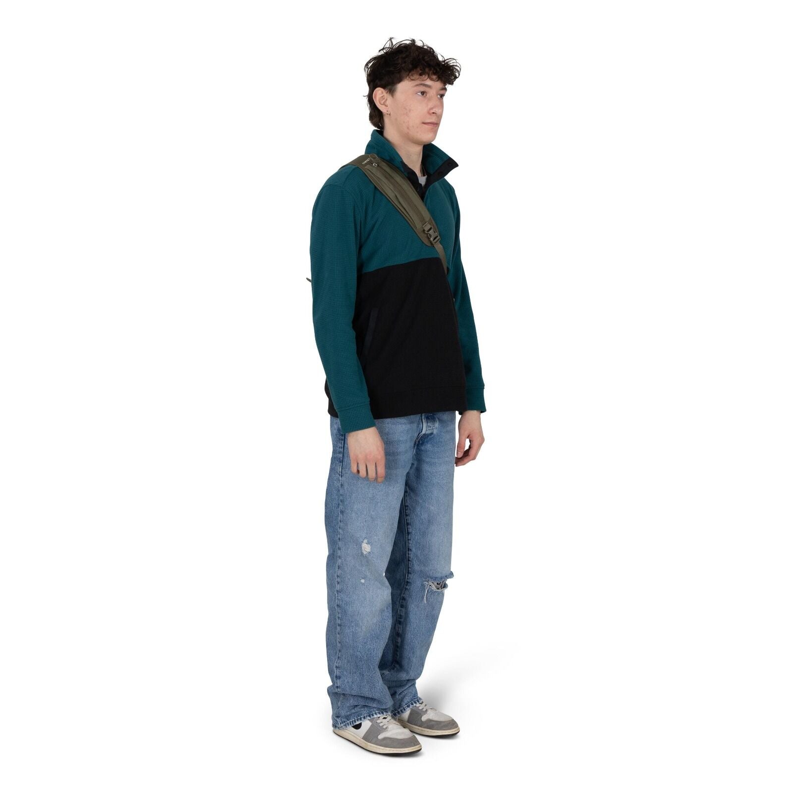A person stands against a white background wearing a casual outfit. They have on a teal and black quarter-zip pullover, light blue baggy jeans with distressed patches, and gray sneakers. An olive-green crossbody sling bag is worn across the chest. The pos