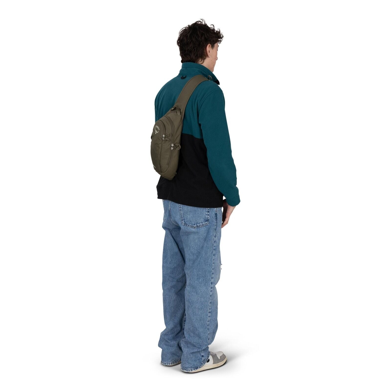 A person is standing turned slightly away, wearing a teal and black fleece jacket with light blue baggy jeans and gray sneakers. They have an olive-green sling backpack worn across the chest, resting on their back; the pack has multiple zippered pockets a
