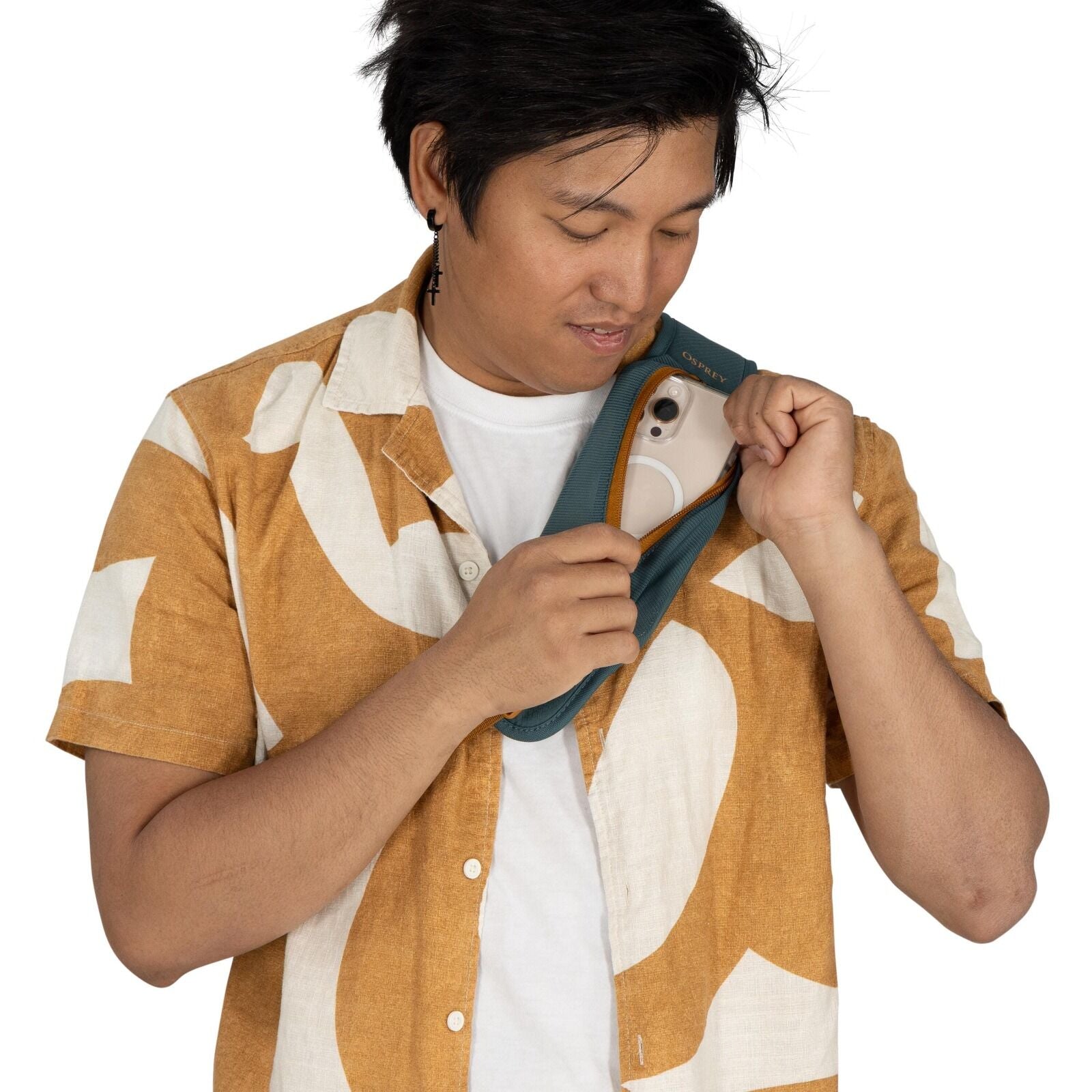 The image shows a person wearing a short-sleeved, button-up shirt with an abstract white pattern on a mustard-colored background, over a white t-shirt. They have a cross-shaped earring in their left ear. The individual is holding a teal-colored bag or sli