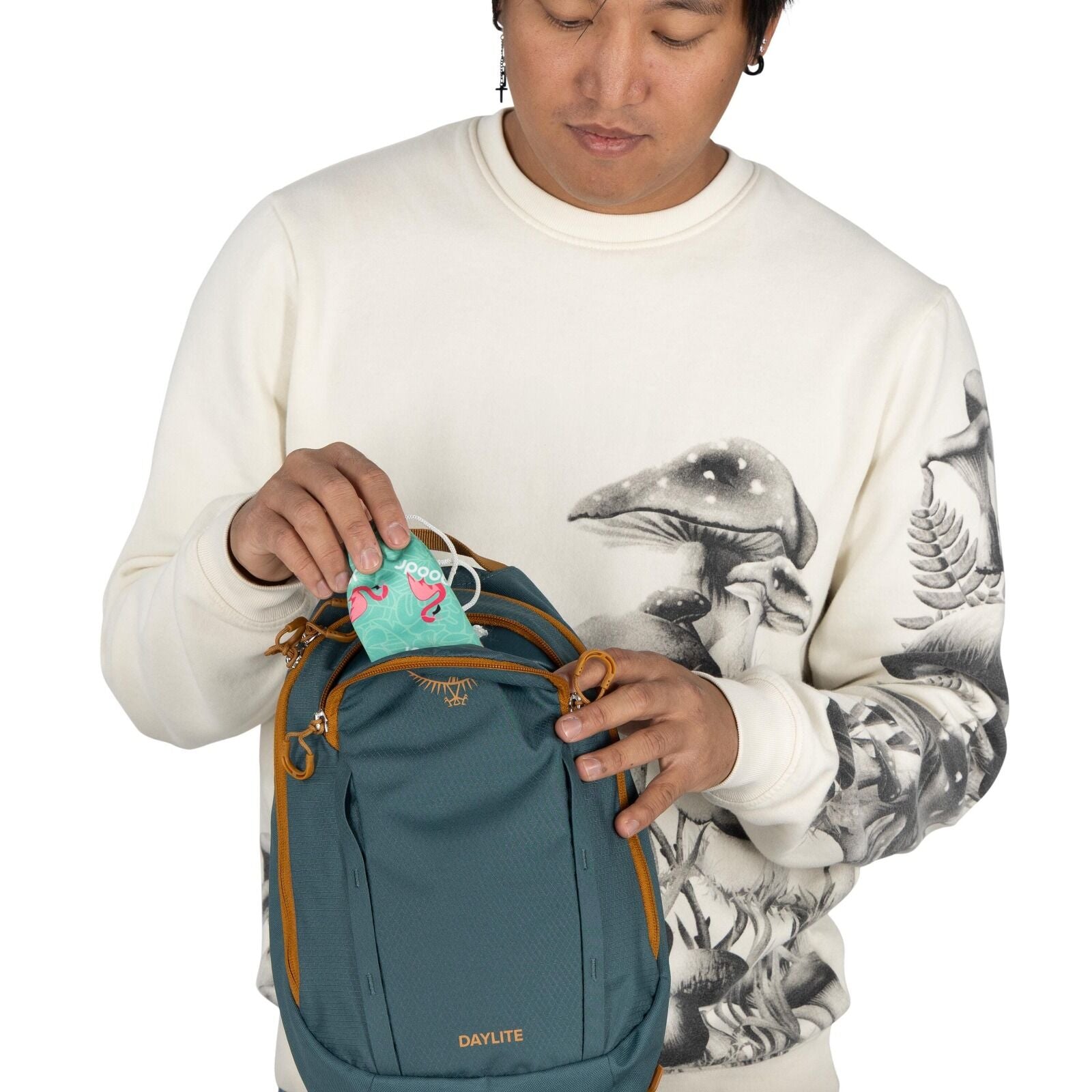 The image shows a person wearing a cream-colored sweatshirt with intricate artwork depicting mushrooms and other foliage in a detailed, monochromatic style. The individual is holding a teal backpack branded with "Daylite" on the front and has tan accents