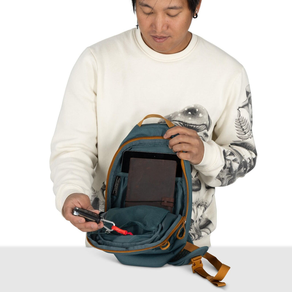 The image shows a person holding an open teal backpack with tan accents, revealing its contents. Inside the backpack, there is a tablet in a dedicated slot, and a mesh pocket containing several items, including a screwdriver. The person is wearing a white