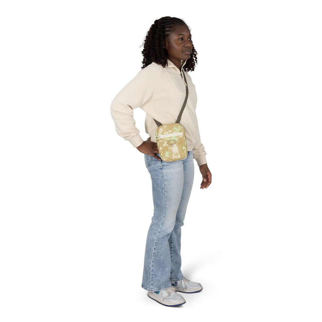 A person stands in a casual pose wearing a cream quarter_zip sweatshirt, light blue flare jeans, and white sneakers with blue accents. They carry a small crossbody bag with a beige and light green floral pattern and olive strap, resting at the hip. One ha