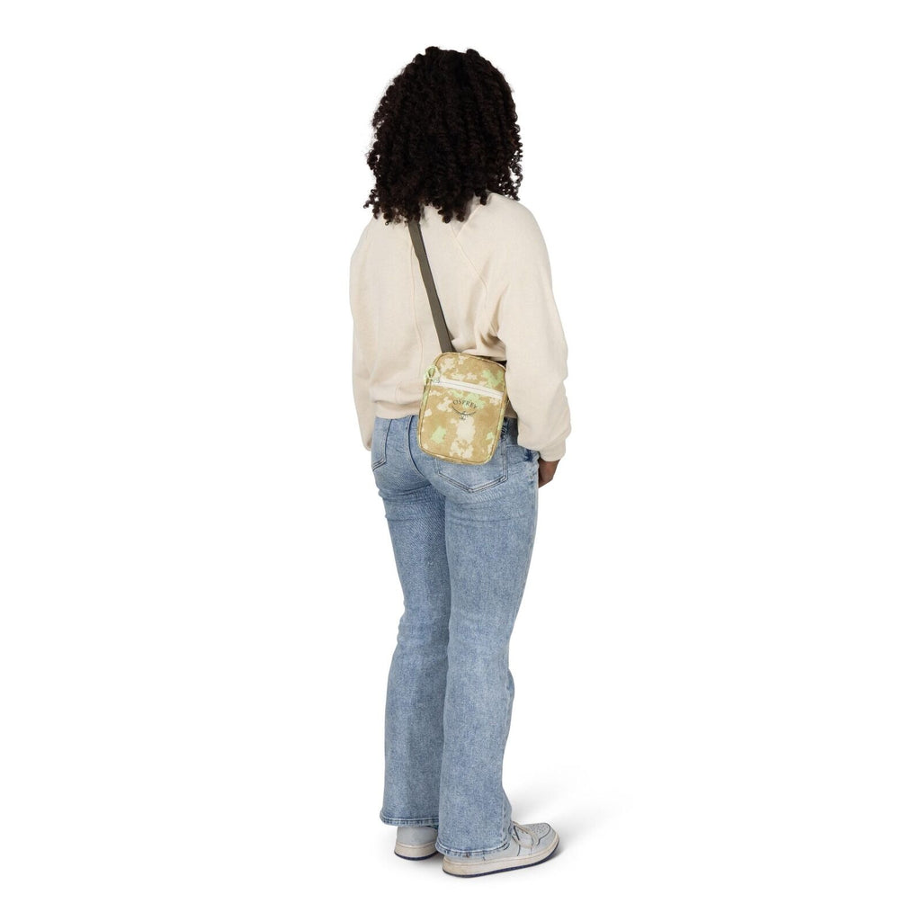 A person is standing with their back slightly turned, wearing a cream sweatshirt, light blue jeans, and gray sneakers. They have a small crossbody bag in a tan camouflage pattern with a dark strap resting on their hip. The background is plain white, keepi