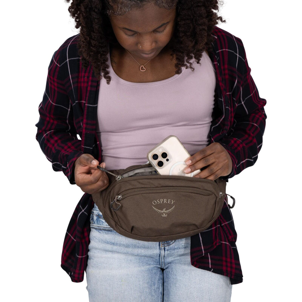 A person is wearing a casual outfit with a lavender tank top, a red-and-black plaid shirt, and light-wash jeans. They have a brown Osprey waist pack strapped around their waist and are placing a smartphone into the main zippered compartment. The Osprey lo