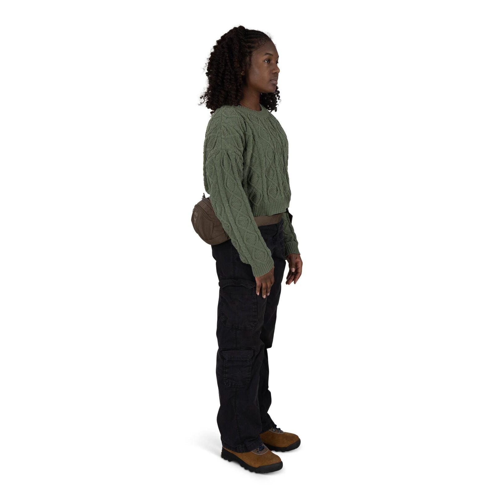 A person stands in profile wearing an olive green cable_knit sweater and loose black cargo pants. They have a small brown crossbody bag resting on their back. The outfit is finished with rugged brown-and-black hiking shoes. The background is plain white,