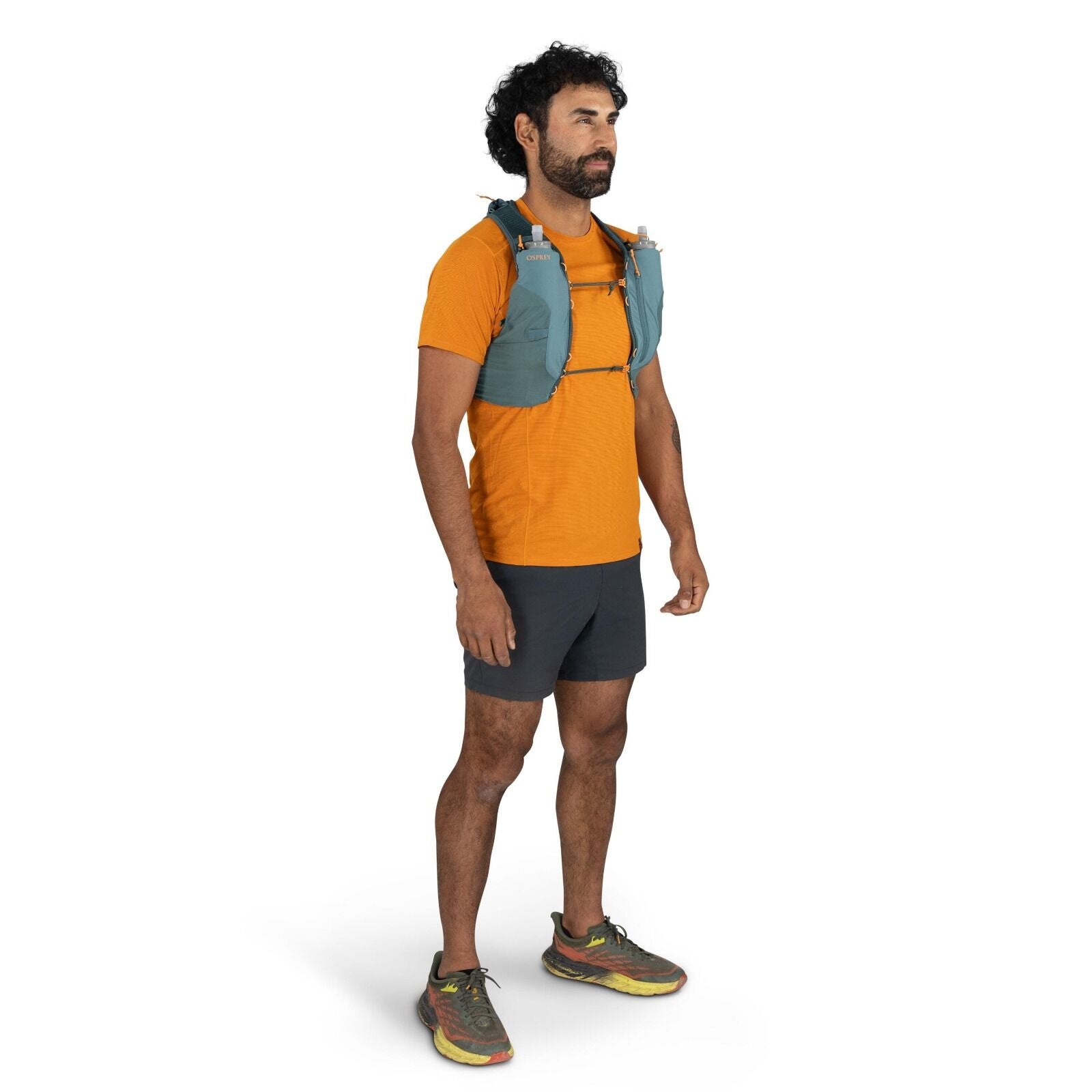 The images show a man standing in a relaxed pose, dressed in athletic gear. He is wearing an orange short-sleeve shirt, black running shorts, and multicolored running shoes. Over his shirt, he has a teal running vest from the brand Camelbak, which is equi