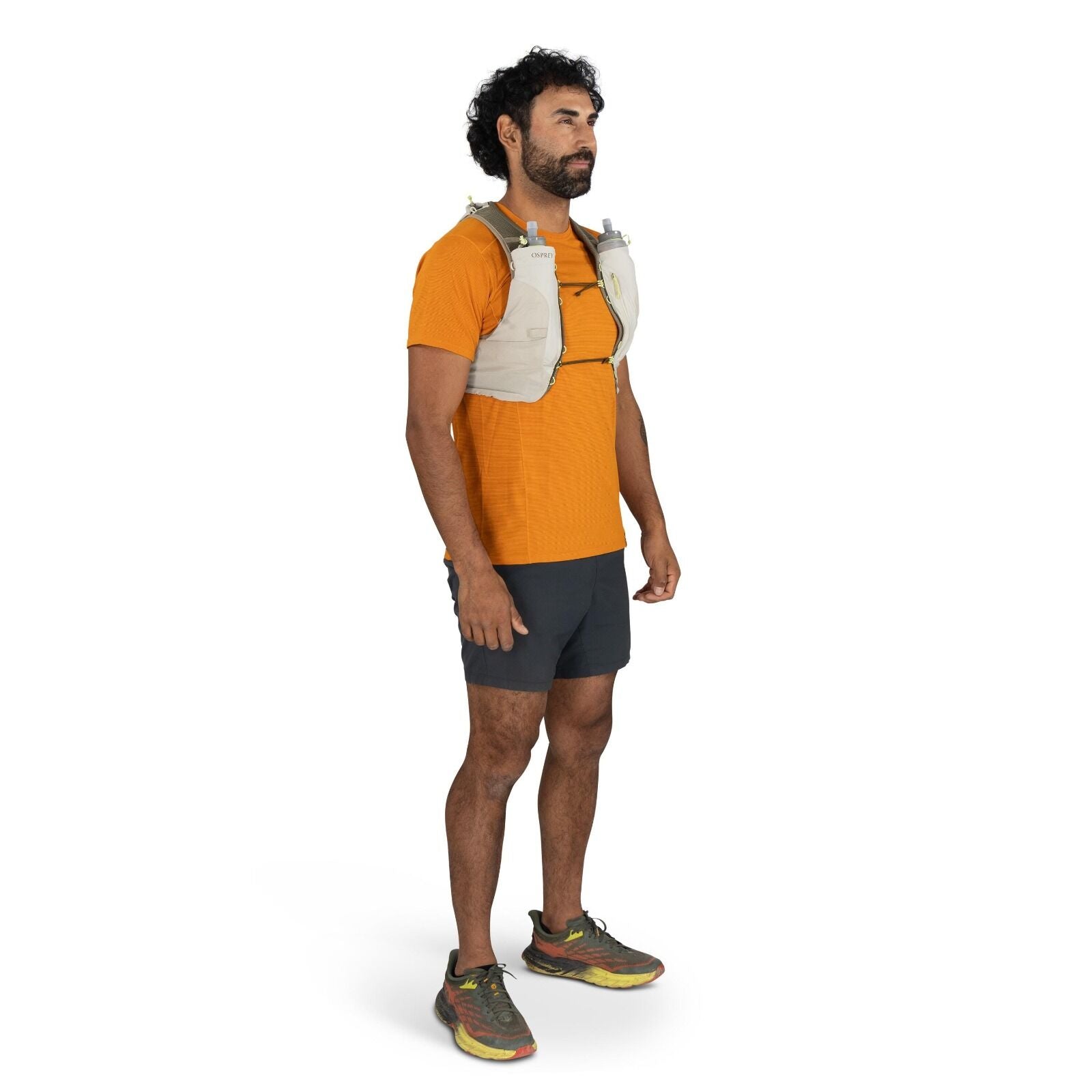 The image shows a person wearing an athletic outfit suitable for running or trail activities. They are dressed in an orange short-sleeved top and dark-colored shorts. The individual is wearing a beige hydration vest with the brand name "Osprey" visible on