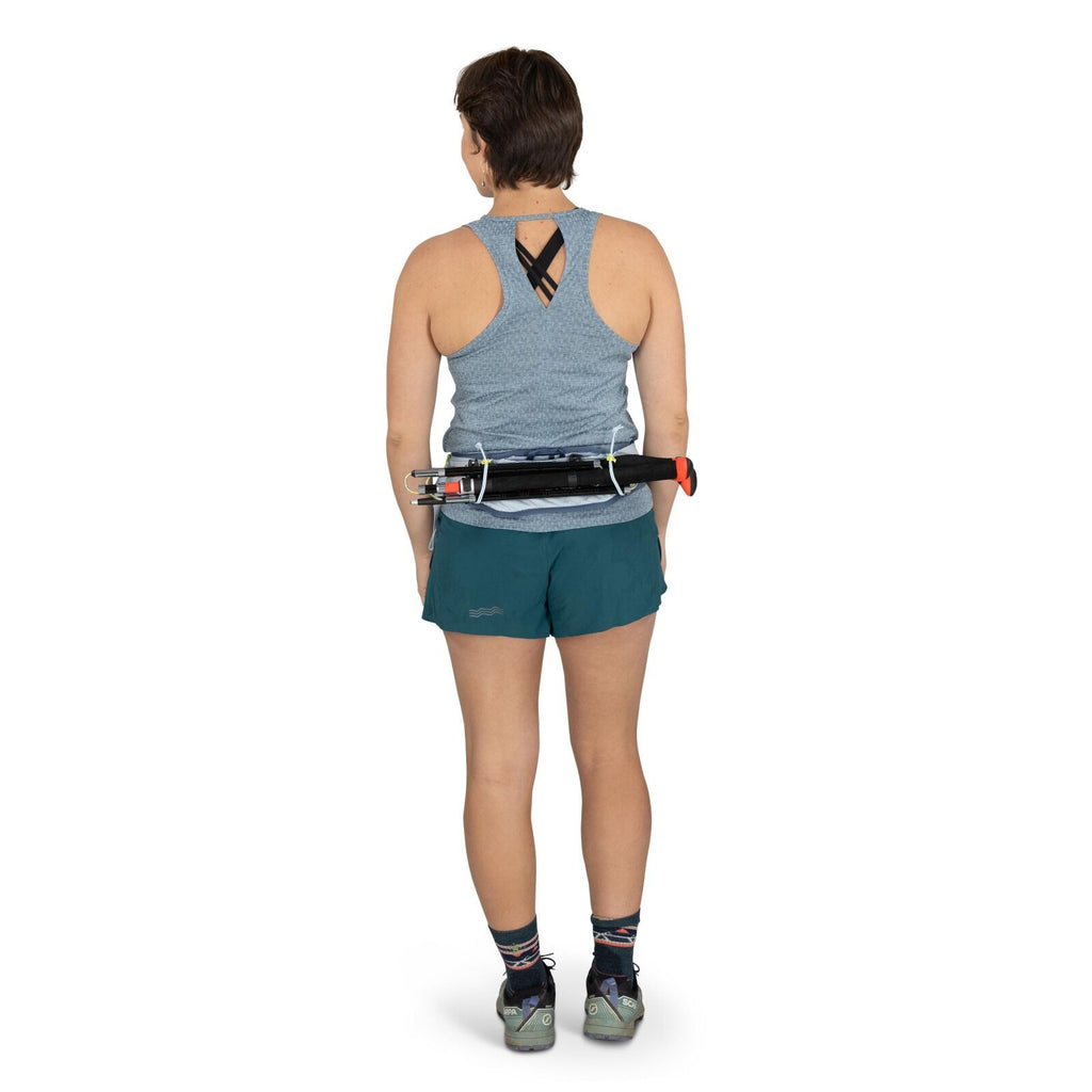 The image shows a woman standing and facing away from the camera, dressed in athletic attire. She is wearing a light blue sleeveless top with a crisscross design at the back and teal shorts with a subtle logo on the side. On her feet are multicolored sock