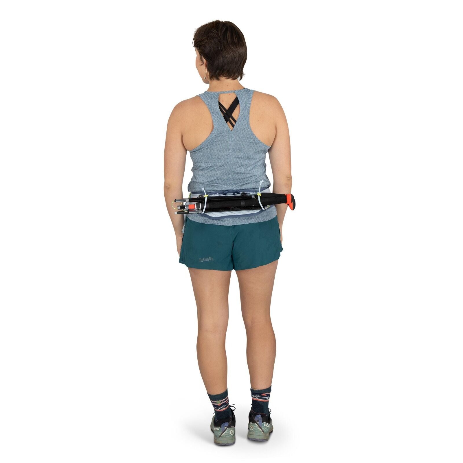 The image shows a woman standing and facing away from the camera, dressed in athletic attire. She is wearing a light blue sleeveless top with a crisscross design at the back and teal shorts with a subtle logo on the side. On her feet are multicolored sock