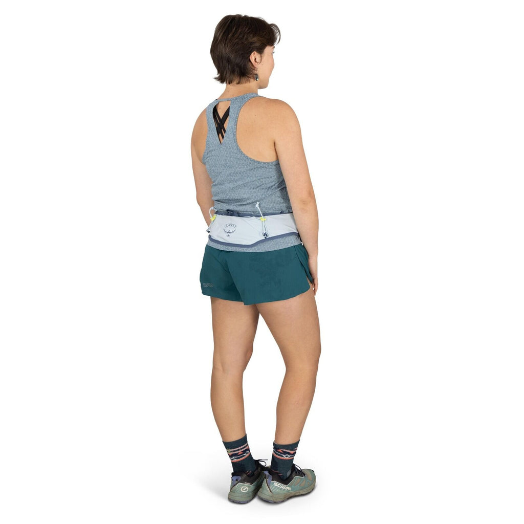 The image depicts a woman standing with her back turned towards the camera. She is wearing a gray, sleeveless, athletic top and dark green running shorts. Around her waist, she has a light gray Osprey brand running waist pack with some adjustable straps.