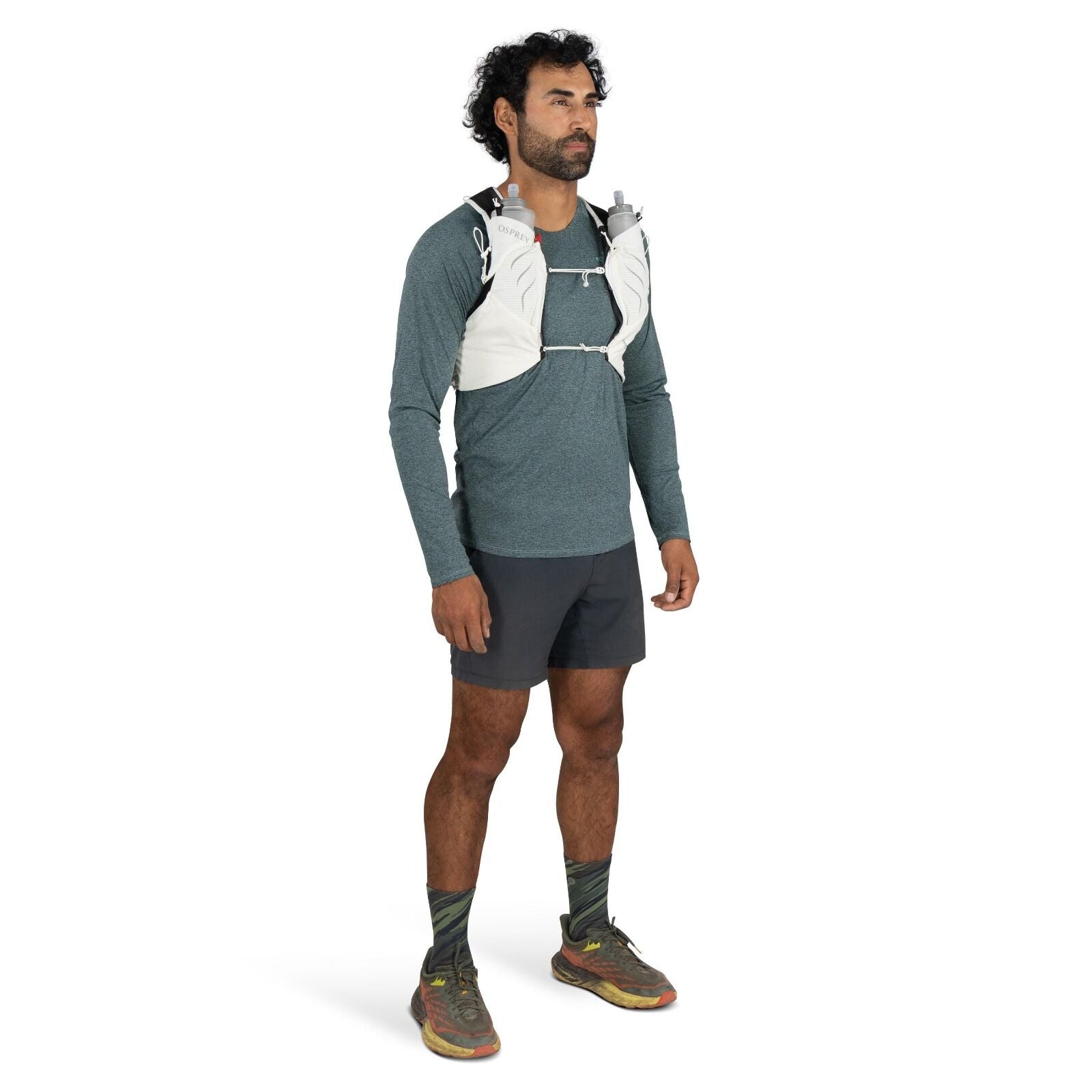 The image shows a man dressed in athletic wear, suitable for running or outdoor activities. He is wearing a long-sleeve teal shirt paired with dark gray shorts. On his feet, he has trail running shoes with a multi-colored pattern and distinctive soles. Th