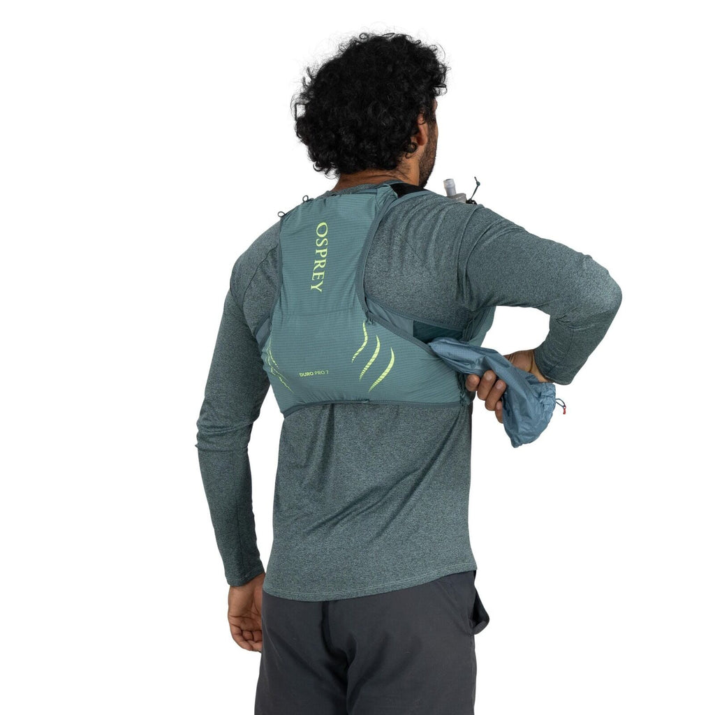 The image shows a man from behind wearing a light green Osprey running vest. The vest is designed with several compartments, and the man is accessing one of them with his left hand, pulling out a light blue item, possibly a jacket or piece of clothing. Th
