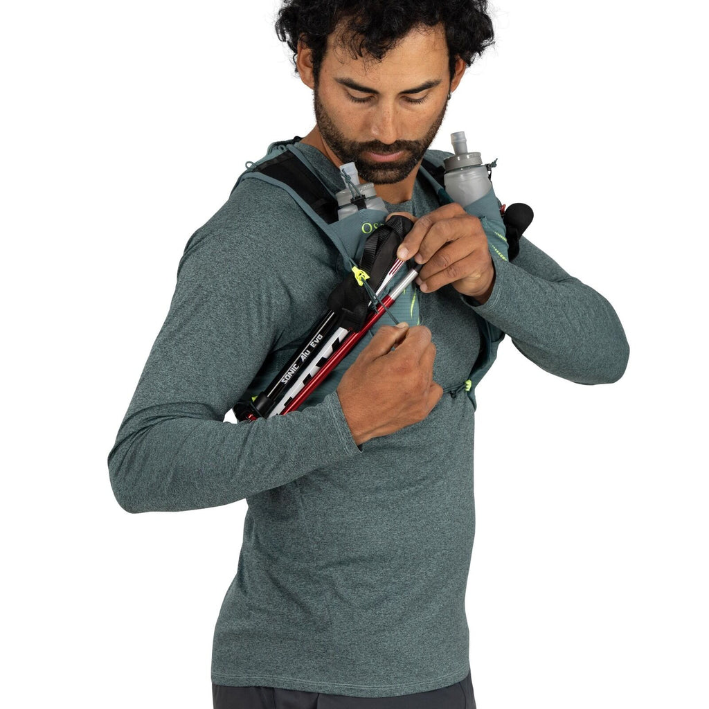 The image shows a person dressed in a teal long-sleeve athletic shirt and dark pants, outfitted with a light blue backpack designed for outdoor activities such as hiking or running. The backpack has shoulder straps and multiple compartments, one of which