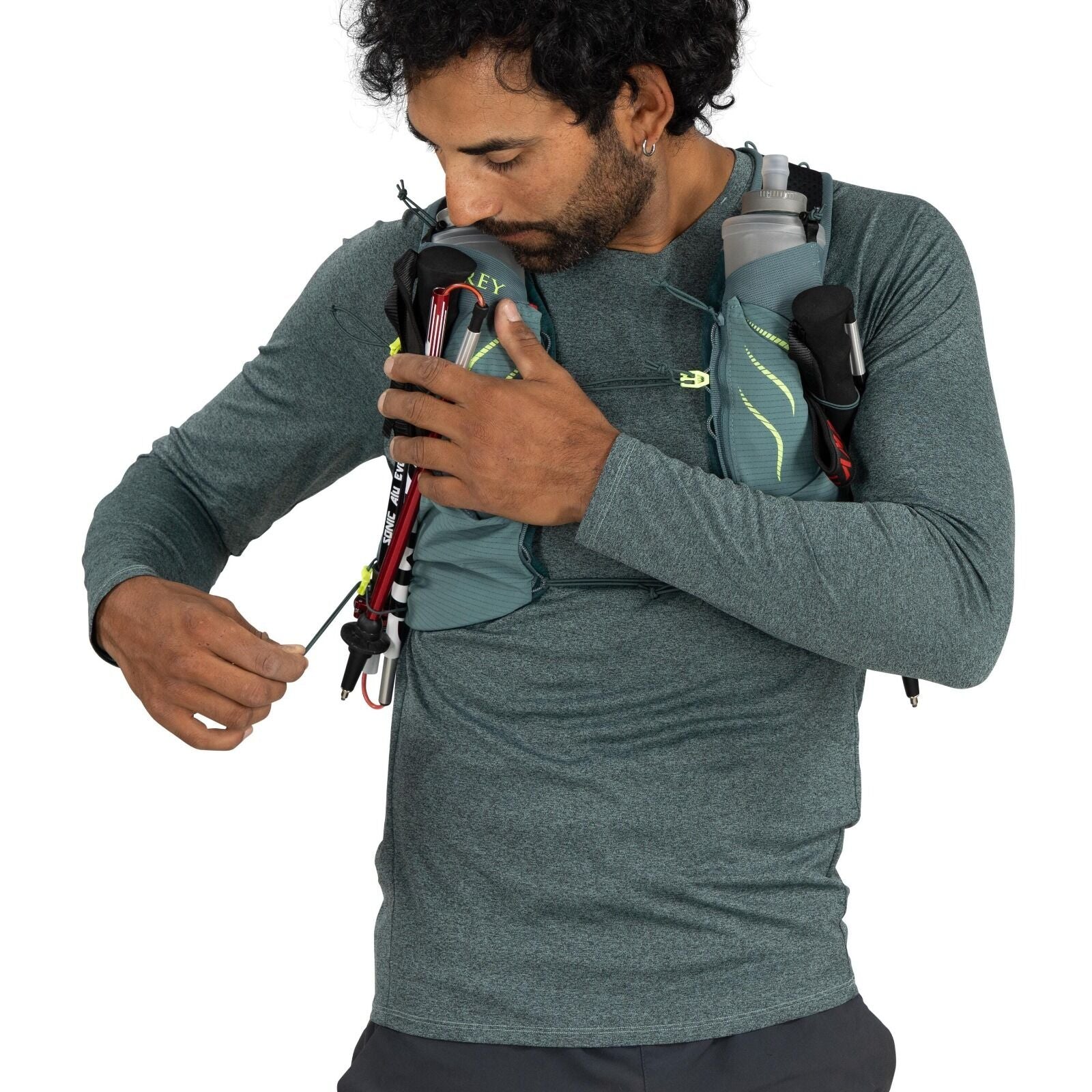 The image shows a person wearing a teal long-sleeve athletic shirt and dark shorts, equipped with a light hiking or running vest. The vest, which is light blue in color, is equipped with various gear including a trekking pole and a water bottle, indicatin
