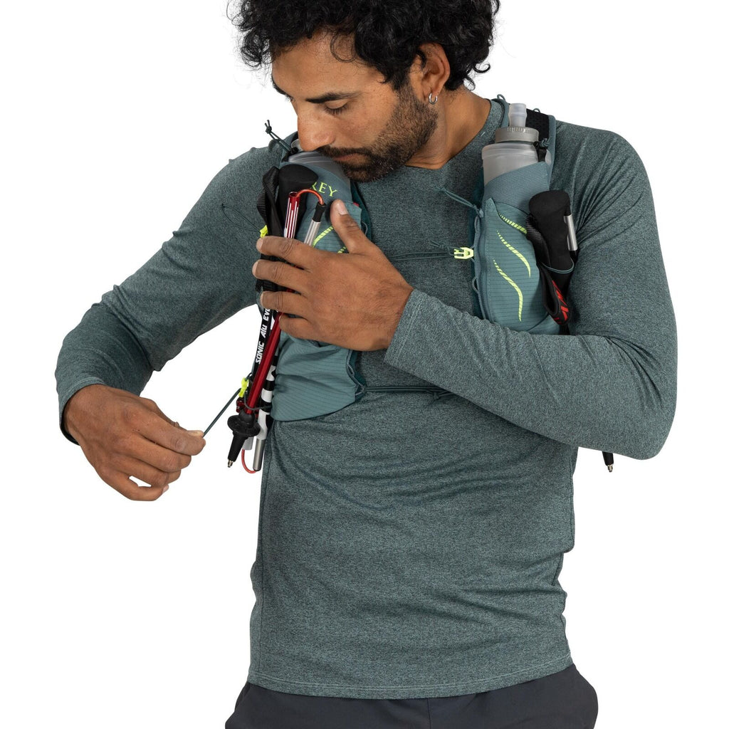 The image shows a person wearing a teal long-sleeve athletic shirt and dark shorts, equipped with a light hiking or running vest. The vest, which is light blue in color, is equipped with various gear including a trekking pole and a water bottle, indicatin