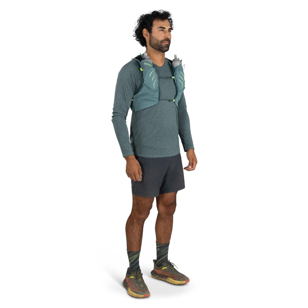 The image features a man standing, dressed in athletic gear suited for outdoor activities, particularly running or hiking. He is wearing a teal long-sleeve shirt paired with dark shorts, and he sports a trail running hydration vest with visible Osprey bra