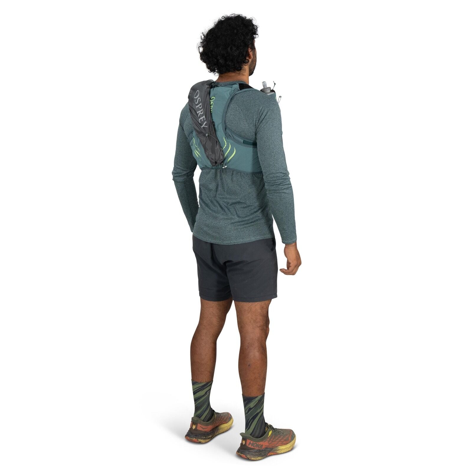 The image shows a man standing with his back to the camera, dressed in outdoor athletic gear. He is wearing a long-sleeved teal shirt, dark gray shorts, and green patterned socks with trail running shoes that have the HOKA logo visible on the back. Over h