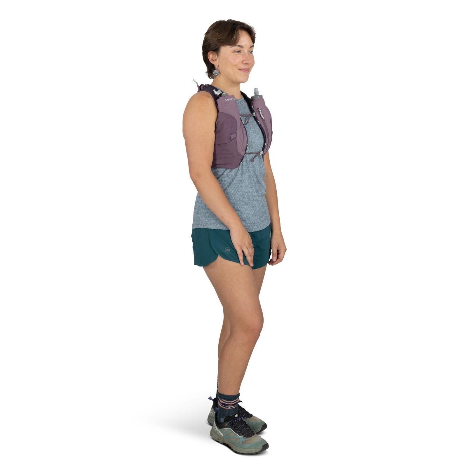 The image shows a person standing in a profile view, wearing outdoor athletic attire. The individual is dressed in a light blue patterned sleeveless top and dark teal shorts, ideal for hiking or running. They are wearing a purple hydration vest with a vis