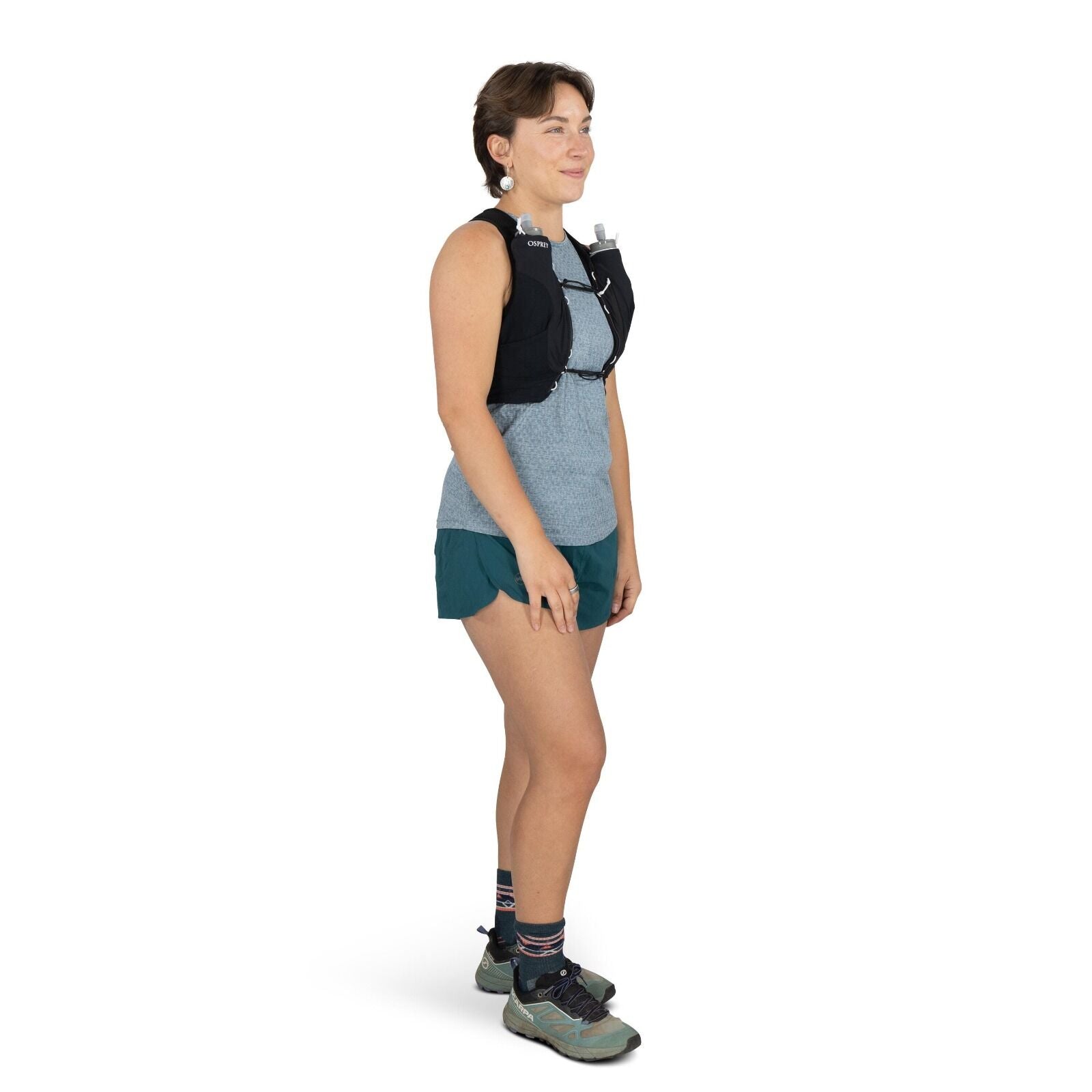 The image shows a person standing sideways to the camera, dressed in outdoor athletic attire suitable for activities like running or hiking. They are wearing a blue-gray patterned sleeveless top and dark teal athletic shorts. The person has a black hydrat